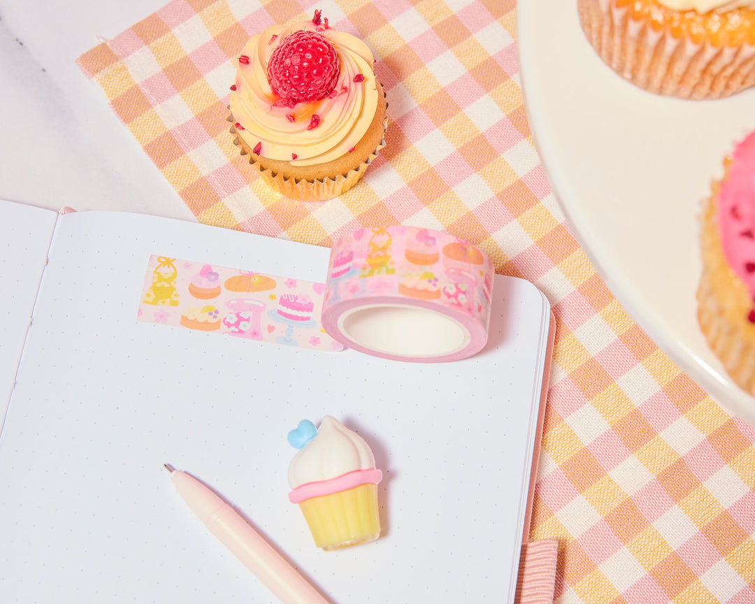 Cupcakes with pink frosting and raspberries on a checkered tablecloth, next to a roll of decorative tape and a cupcake-shaped gel pen.