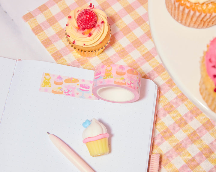 Cupcakes with pink frosting and raspberries on a checkered tablecloth, next to a roll of decorative tape and a cupcake-shaped gel pen.