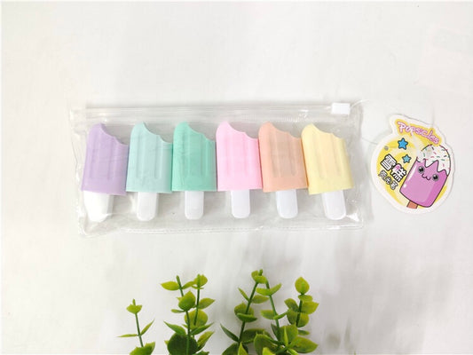 set of 6 popsicle highlighters in purple, blue, green, pink, orange and yellow, inside a clear bag lying on a white background with a green plant underneath