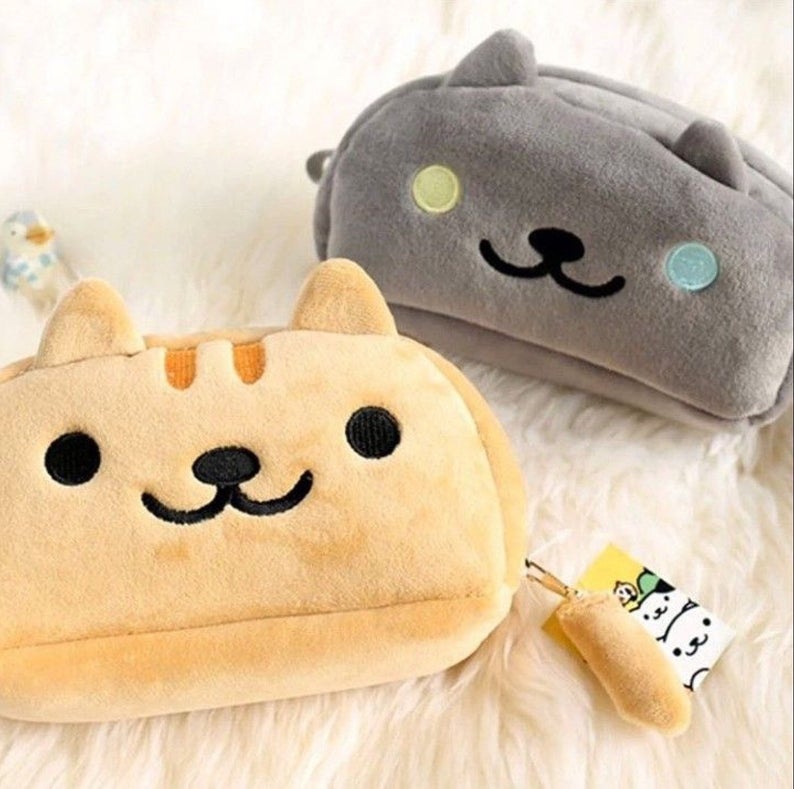 2 cat plush pencil cases featuring faces from the Neko Atsume game. The bottom is ginger and the top is grey with one yellow and one blue eye