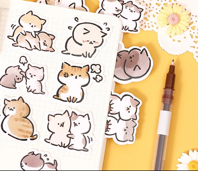 7 cat stickers laid onto a grid notebook, there are 4 more cat stickers coming out from the other pages. The book is laid on a yellow background with flowers and a gel pen